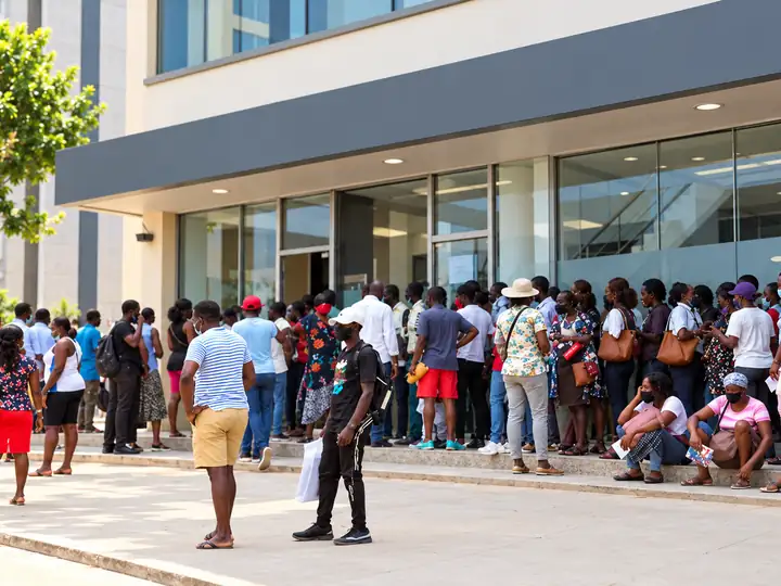 Fila longa de pessoas em pé à entrada envidraçada de um edifício de atendimento.